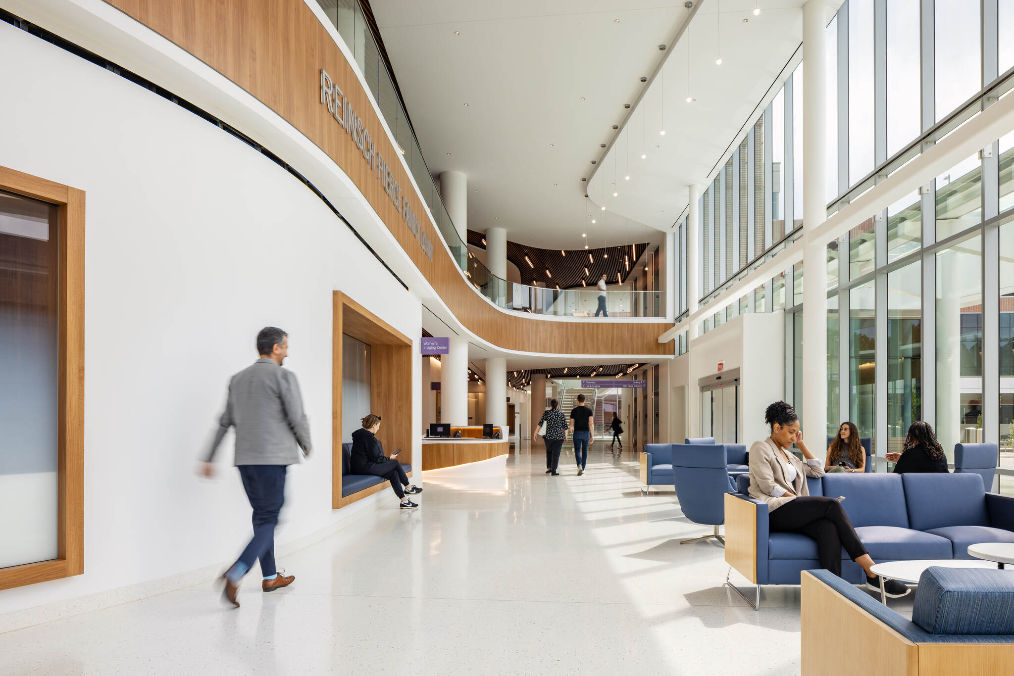 Bright modern hospital lobby with people walking and sitting on blue sofas near large windows.