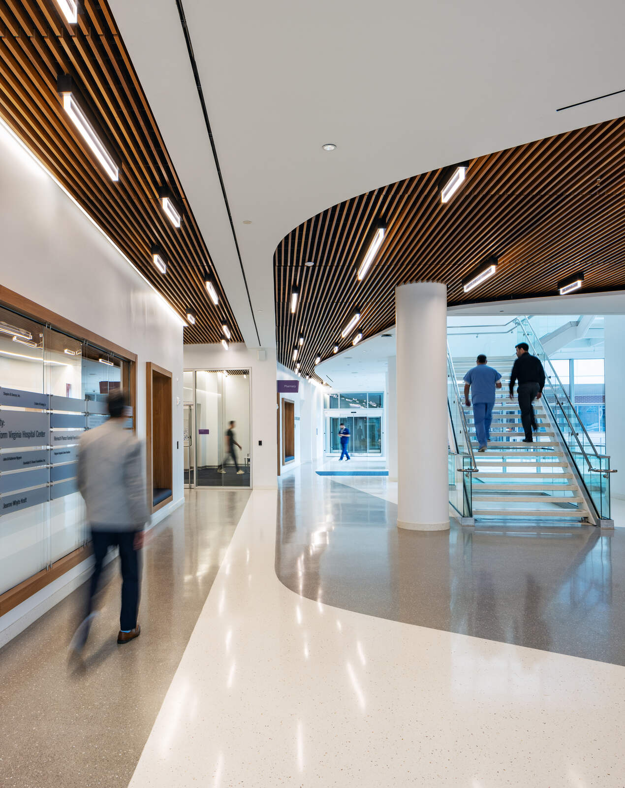 Modern hospital corridor with wooden ceiling panels, glass stairs, and blurred people walking and ascending stairs.