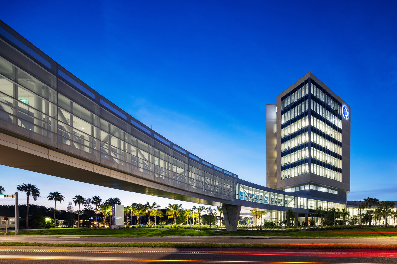 Modern glass building connected to a skybridge with palm trees and a twilight sky background.