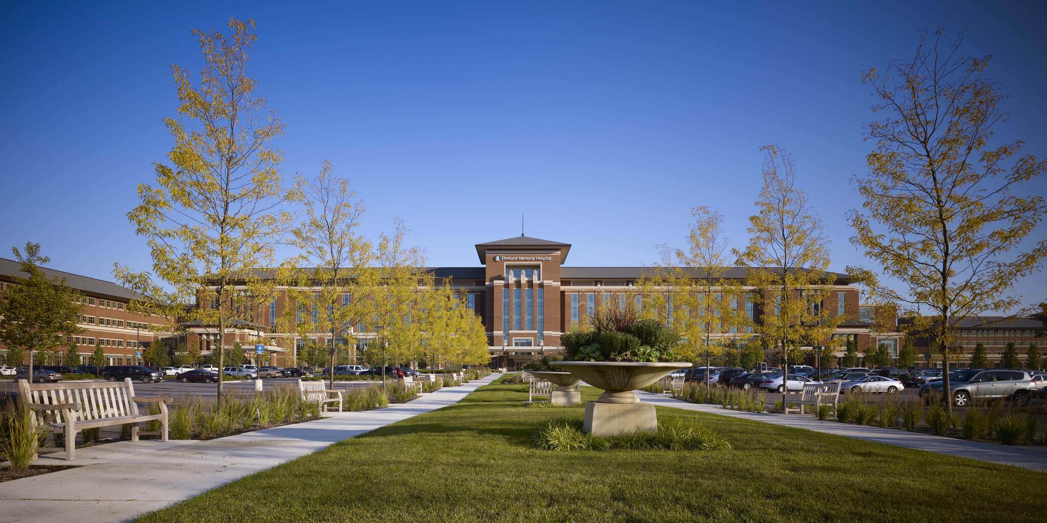 Wide front view of Elmhurst Memorial Hospital with pathways, benches, trees, and blue sky.