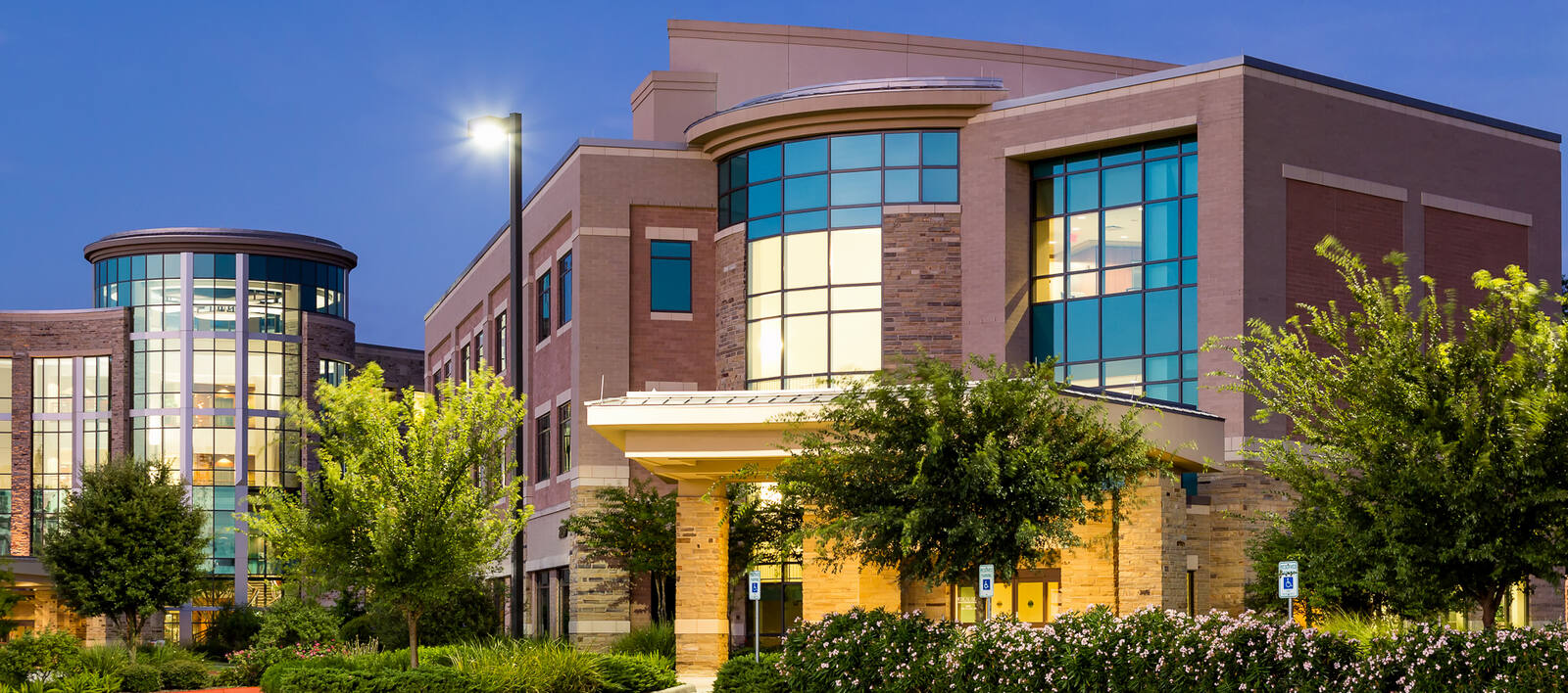 Modern hospital building with large windows, green landscaping, and a lit street lamp at twilight.