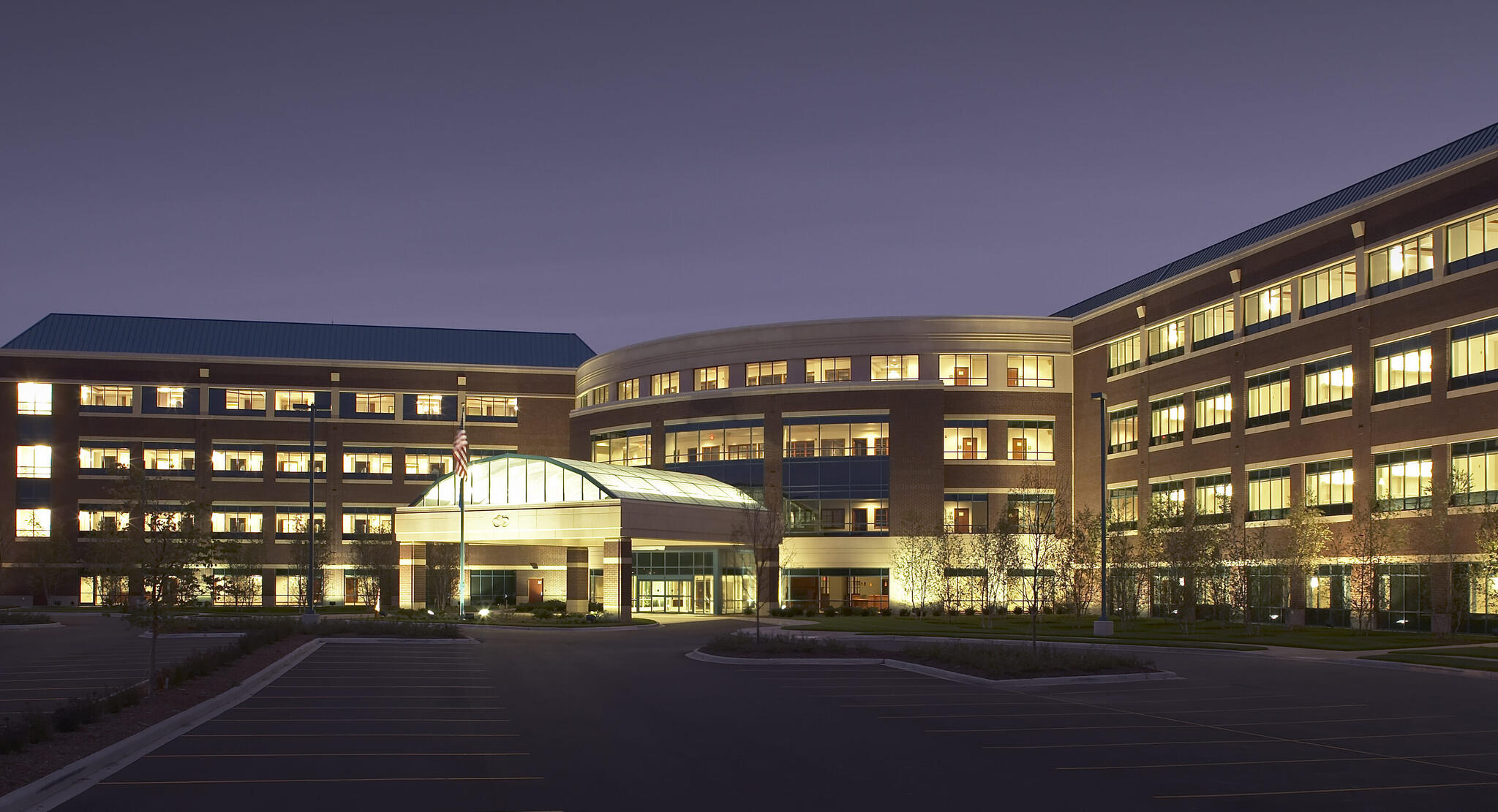 Large illuminated medical center building at dusk with empty parking lot and American flag.