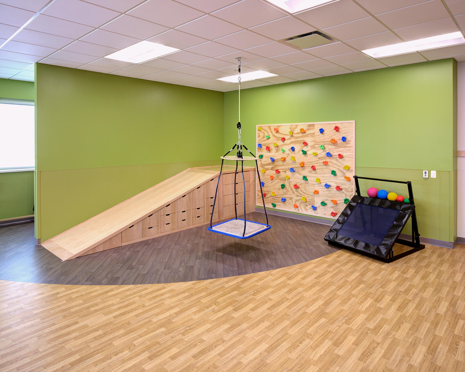 Therapy room with a wooden ramp, hanging swing, climbing wall, and ball return game setup.