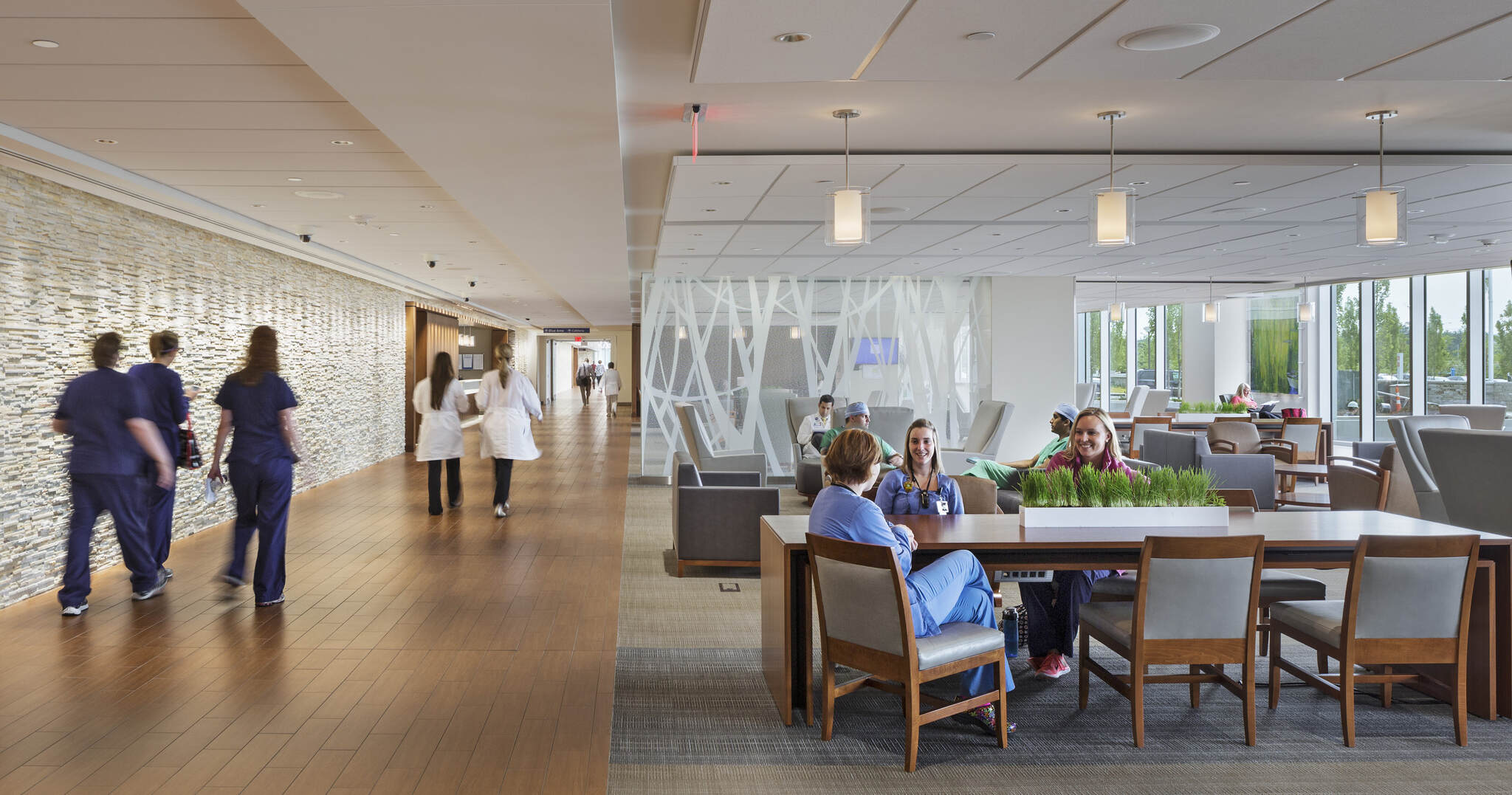 Hospital staff walking in a bright corridor and others sitting and talking in a modern lounge area.