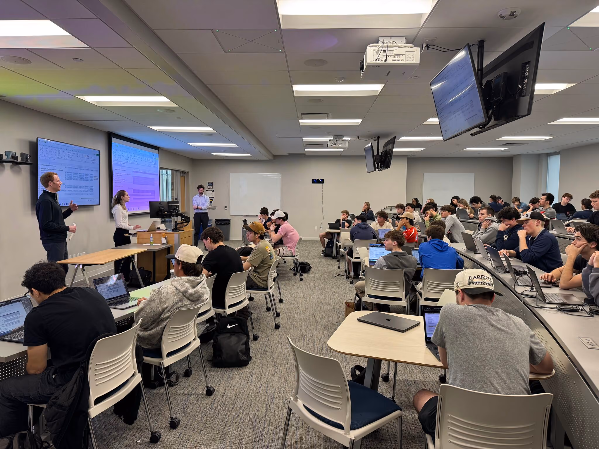 A classroom with students working on laptops and three instructors presenting at the front with screens.