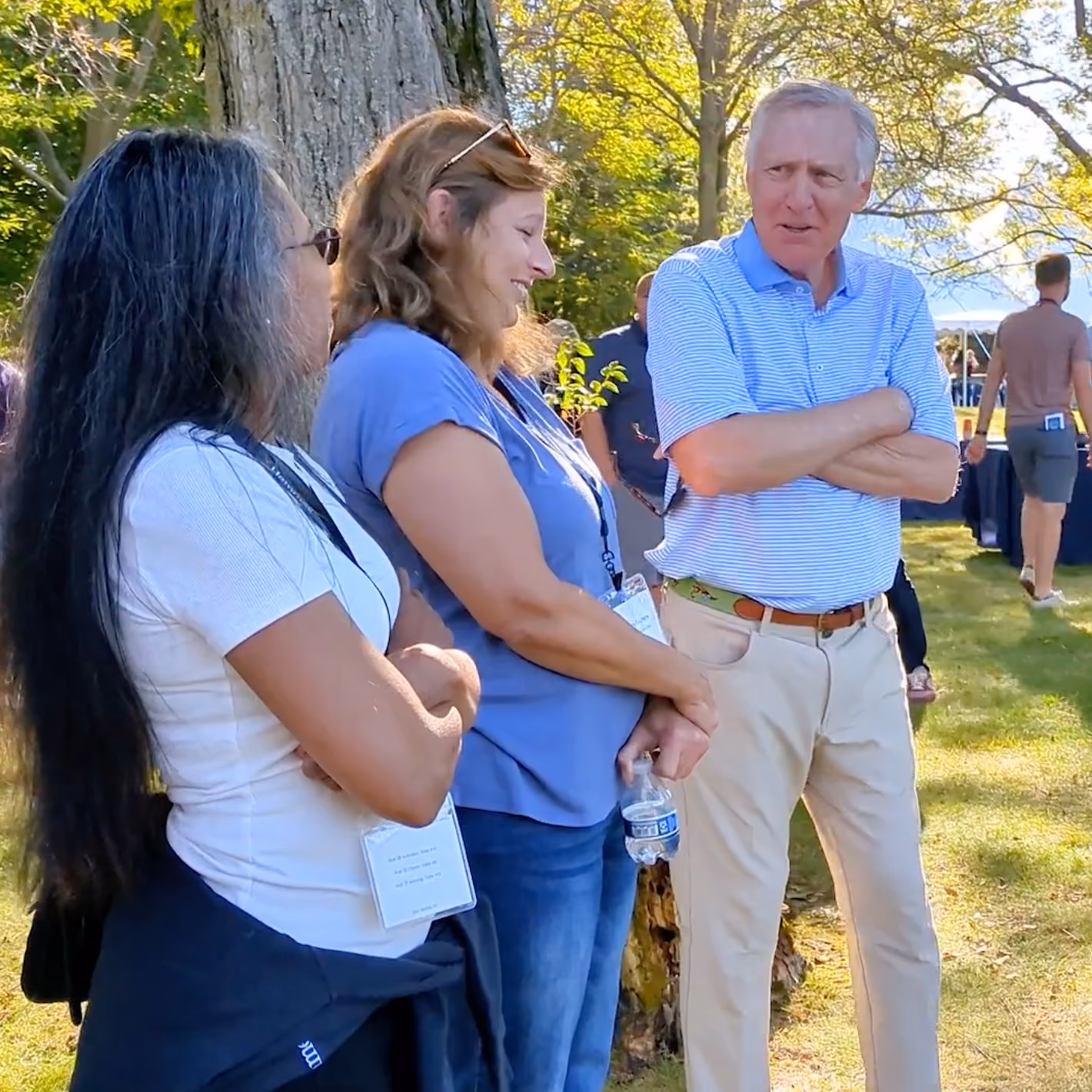 Three people standing outdoors, two women and one man, engaged in conversation by a tree.