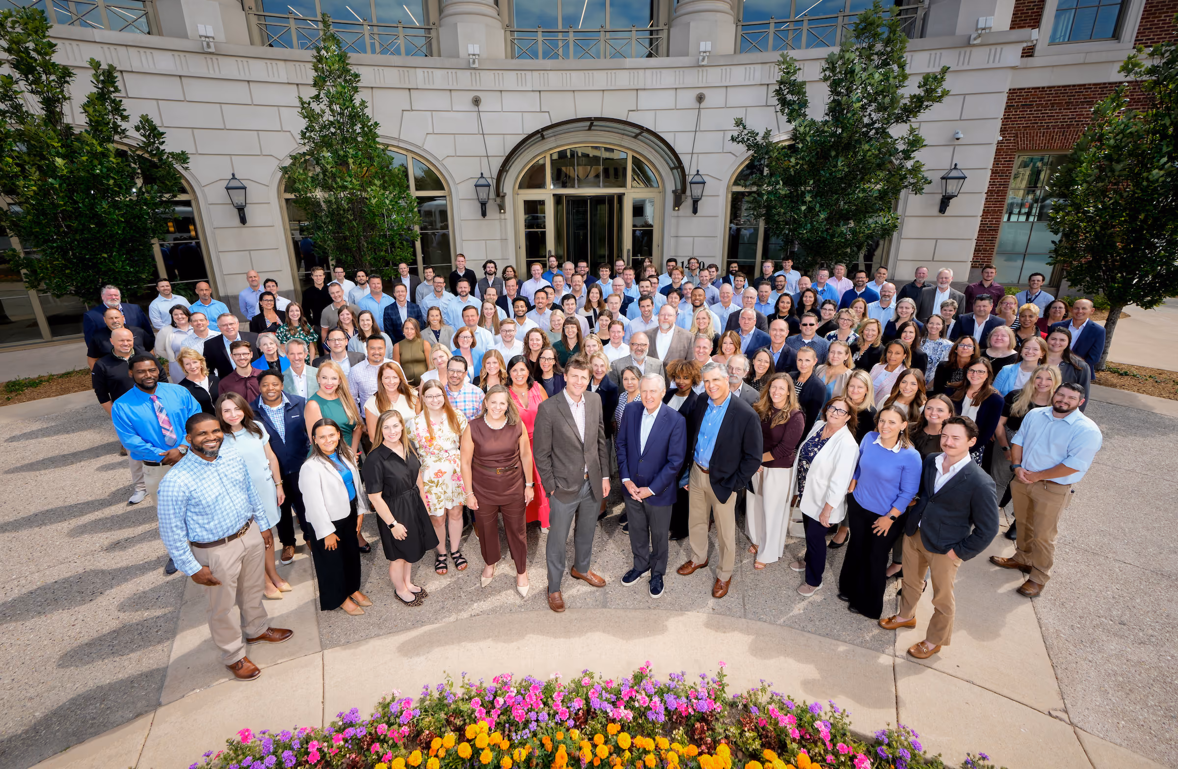Large diverse group of Hammes Employees posing outside a building with flower bed in front.