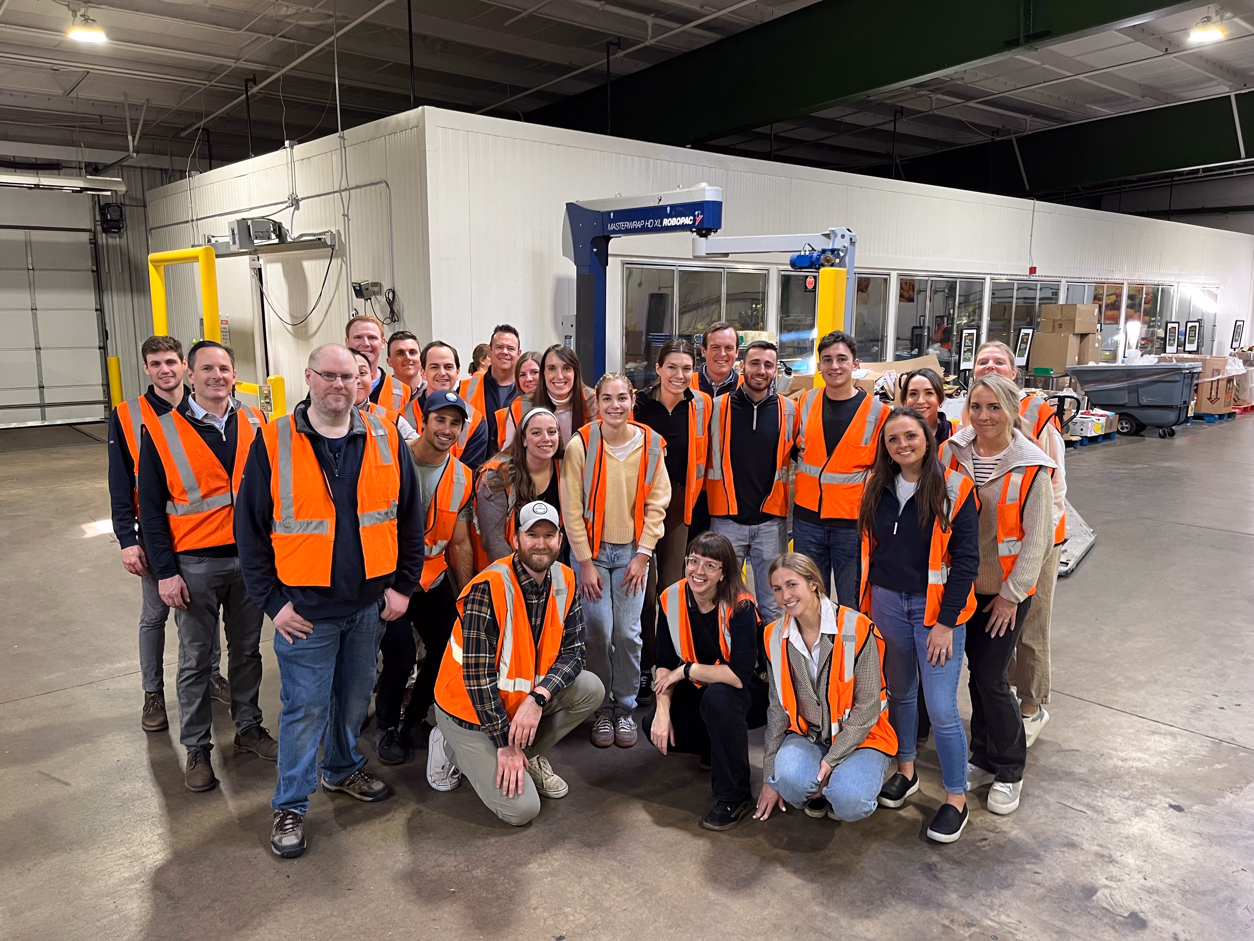 Group of people wearing orange safety vests posing inside a spacious warehouse facility.