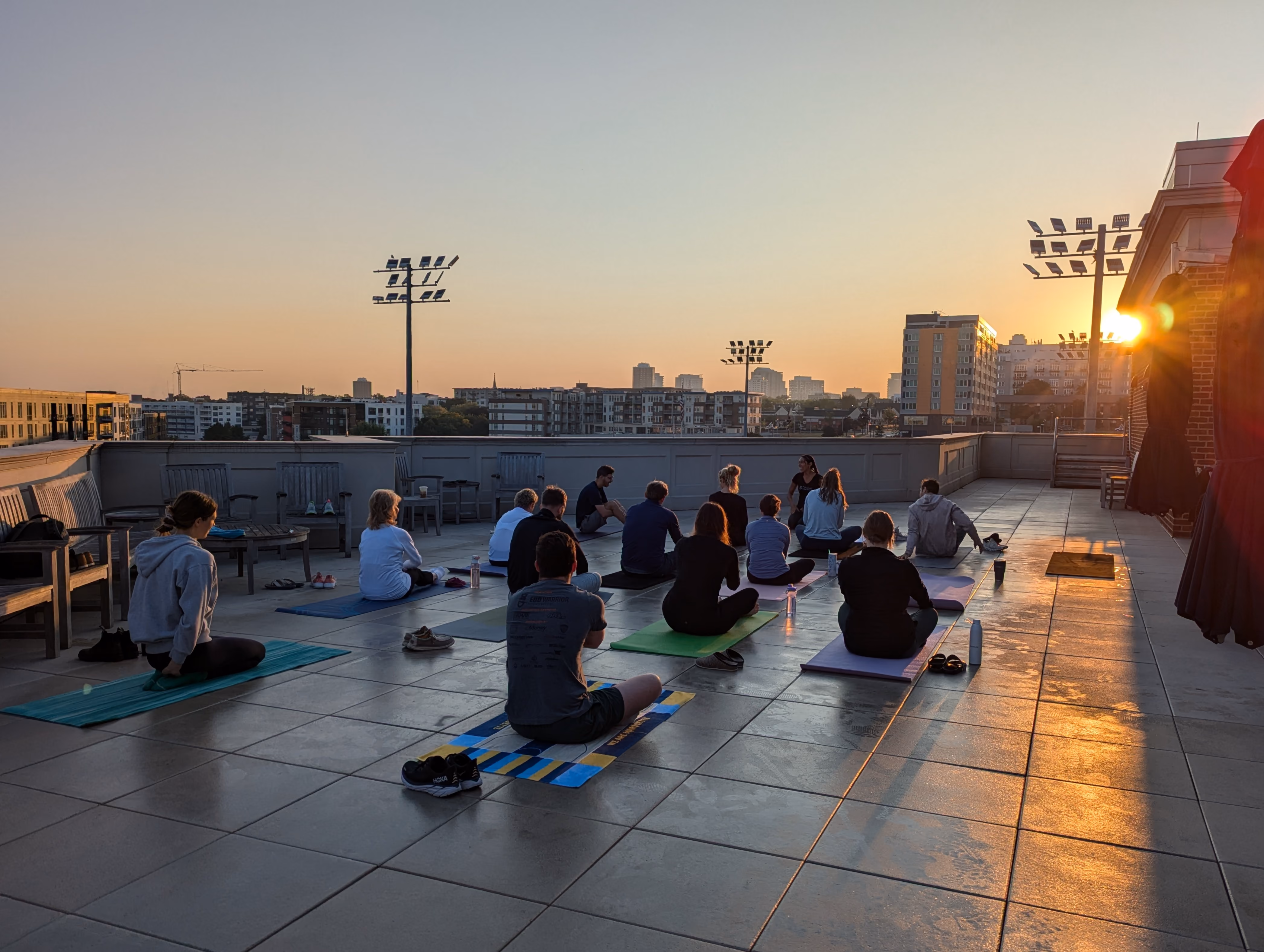 Group of people sitting on yoga mats on a rooftop at sunset with city buildings in the background.