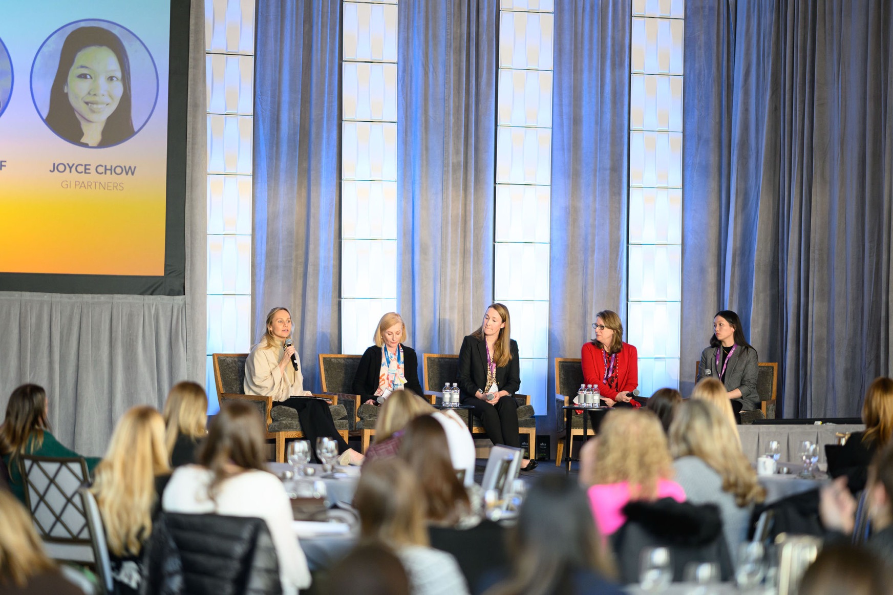 Panel of five women seated on stage discussing at a conference with audience in foreground.
