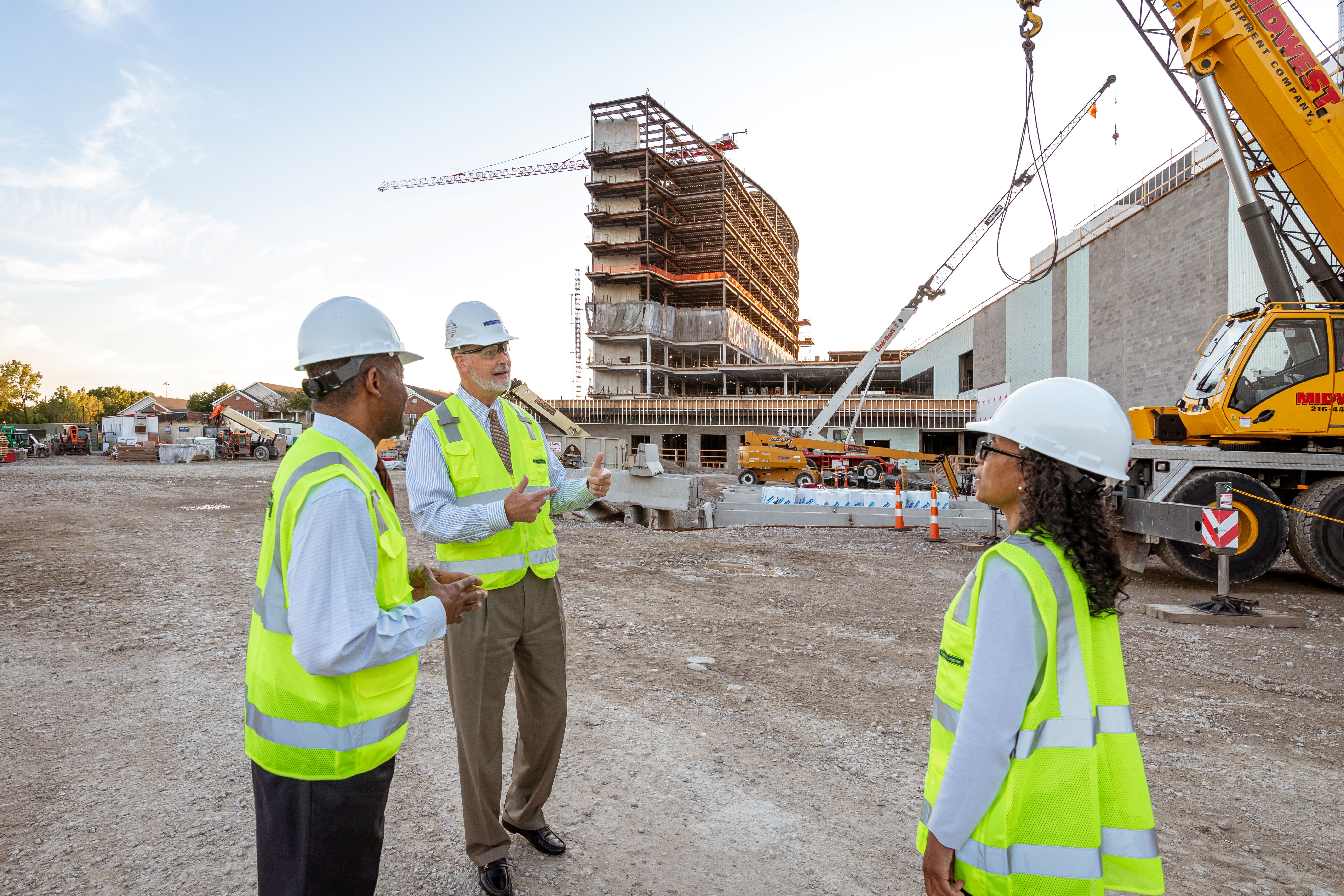 Three construction workers wearing hard hats and yellow vests discussing at a building site.