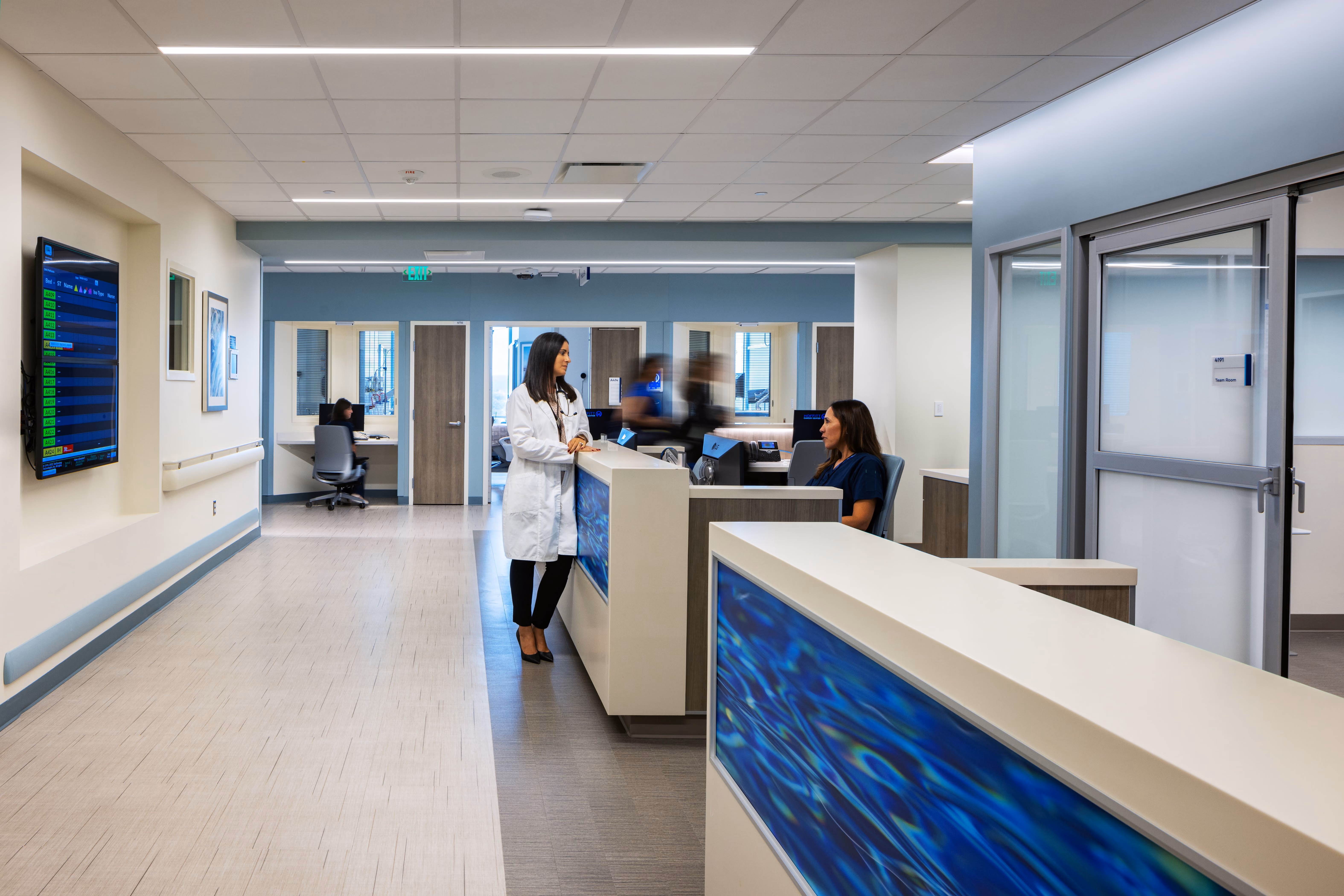 A doctor in white coat talks to a seated nurse at a hospital nursing station with staff and monitors nearby.