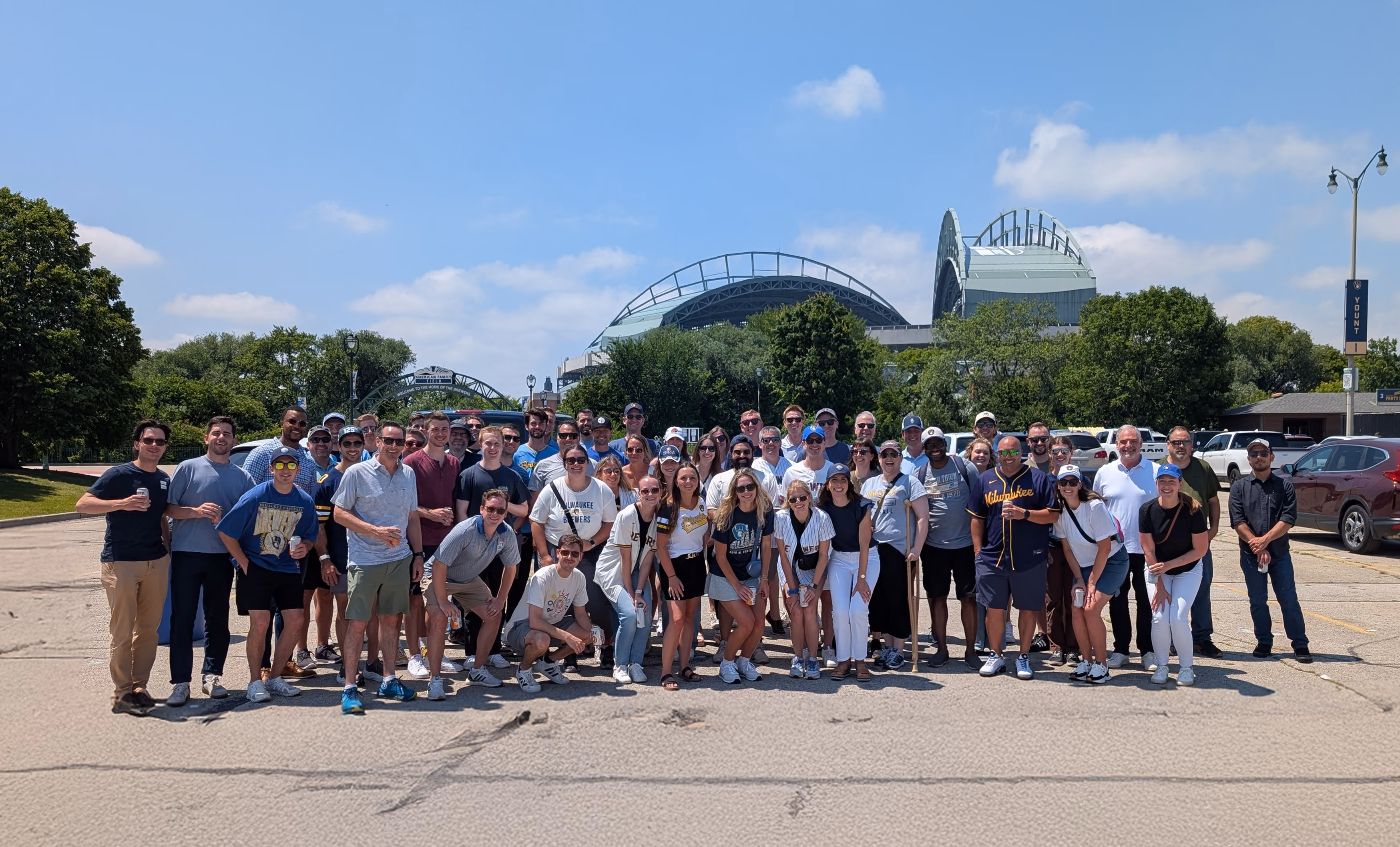 Large group of Hammes Teammates on a paved area outdoors on a sunny day, with trees, cars, and Brewer's stadium visible in the background under a blue sky with few clouds.