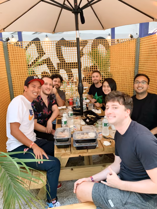 Group of six friends sitting around a wooden picnic table under a large umbrella, smiling with takeout food containers and drinks on the table.
