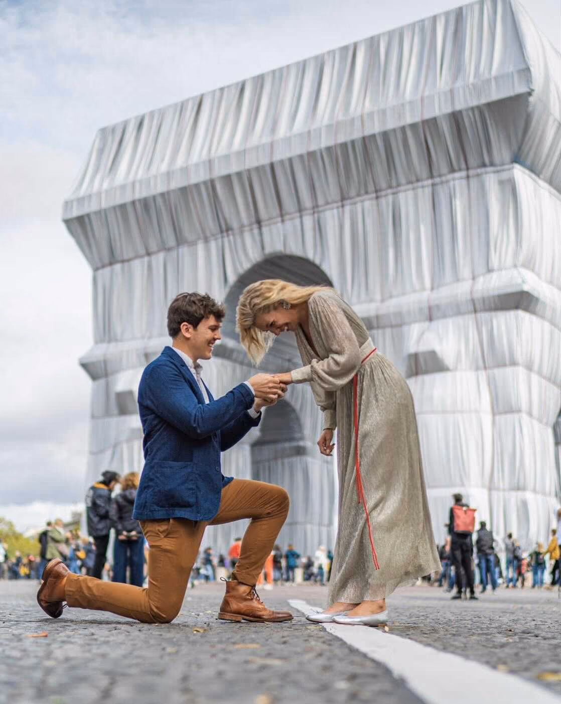 Man proposing on one knee to woman in front of the wrapped Arc de Triomphe with people in the background.