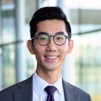 Smiling man wearing glasses, a suit, and a tie in a modern office setting.