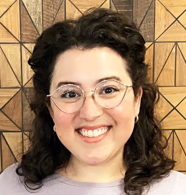 Smiling woman with curly dark hair, round glasses, and a light purple top in front of a wooden geometric patterned wall.
