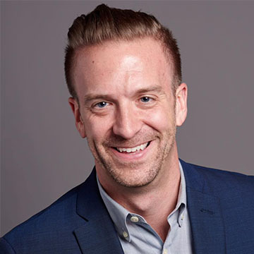Smiling man with short brown hair wearing a blue blazer and light gray shirt against a neutral background.
