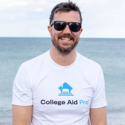 Smiling man wearing sunglasses and a white College Aid Pro t-shirt standing by the ocean.