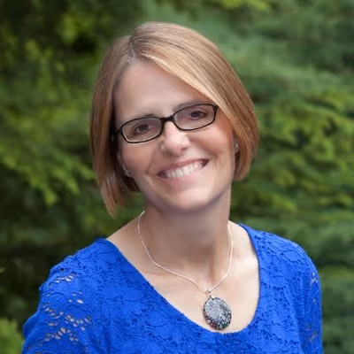 Smiling woman with short blonde hair, glasses, and a blue lace top wearing a round pendant necklace standing outdoors with green foliage background.
