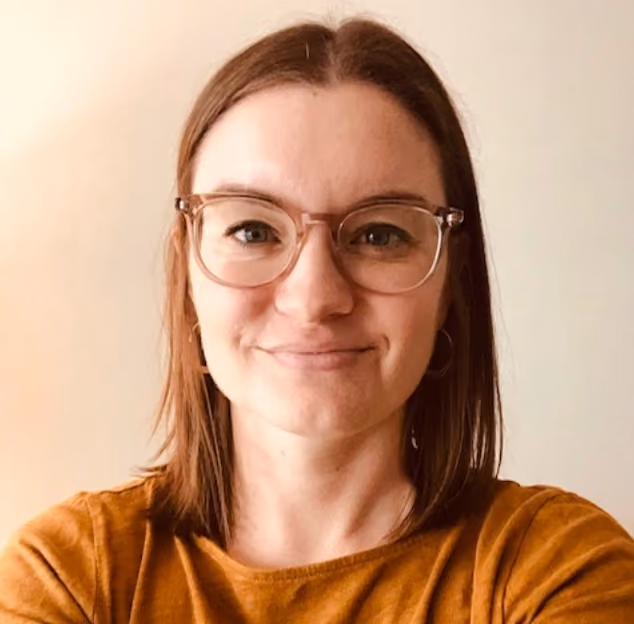 Smiling woman with shoulder-length brown hair wearing round glasses and a mustard-colored shirt against a light background.