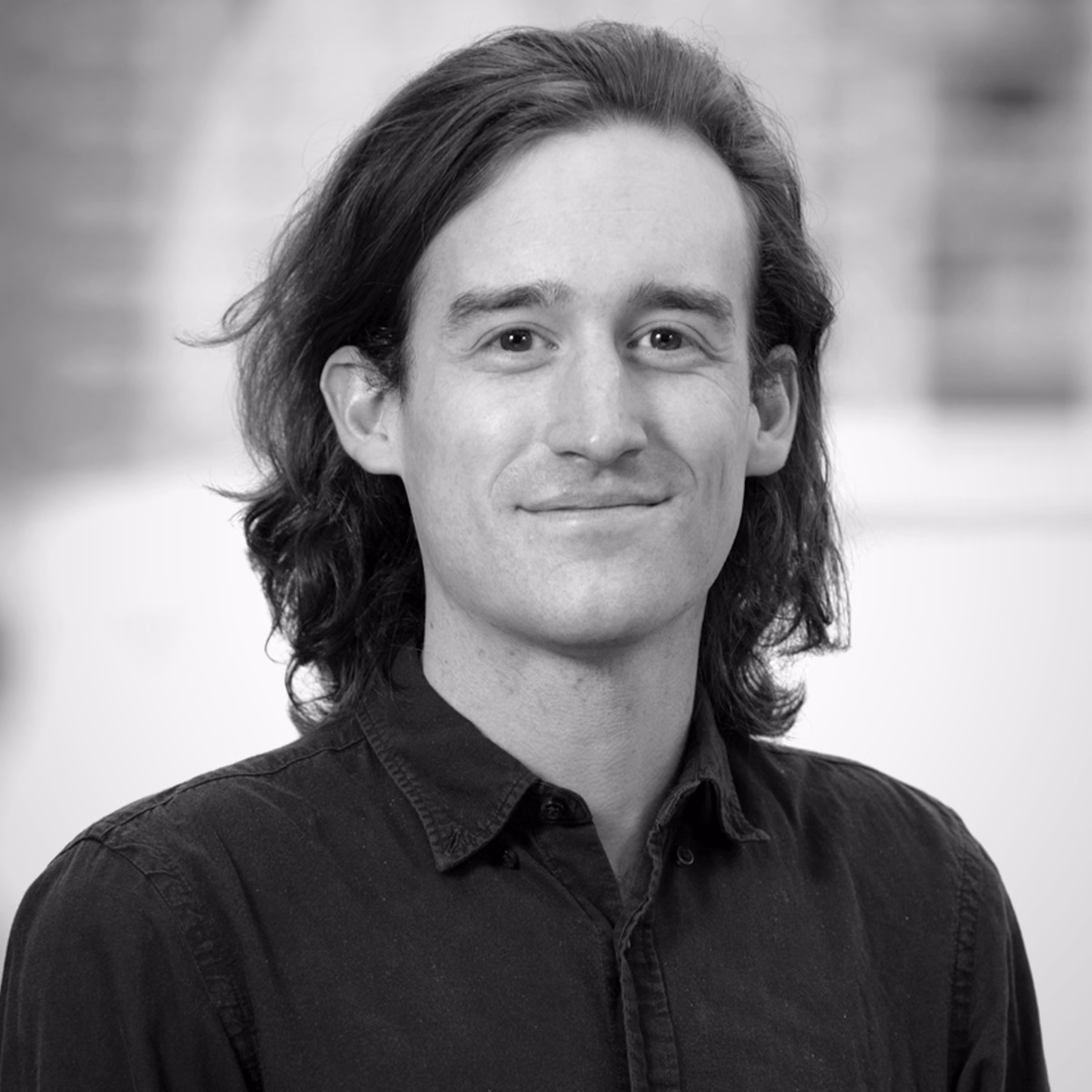 Black and white portrait of a man with shoulder-length hair wearing a dark button-up shirt, smiling slightly.