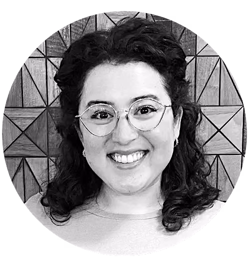 Smiling woman with curly dark hair, wearing round glasses and earrings, against a geometric wooden background.