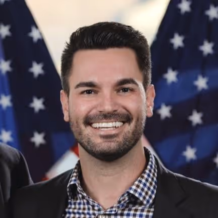 Smiling man with dark hair and beard wearing a checkered shirt and blazer in front of American flags.