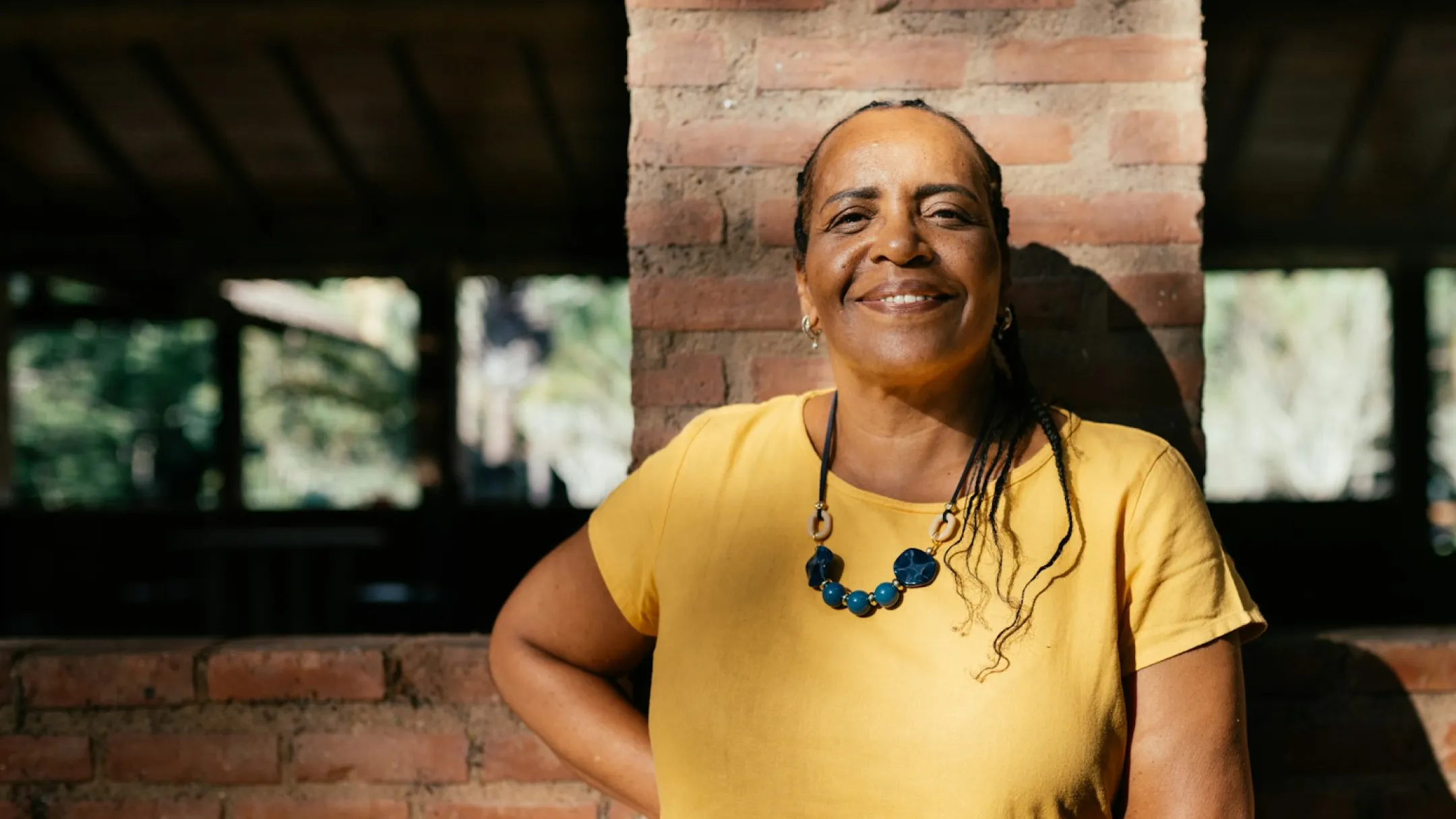 Smiling woman in a yellow shirt and blue necklace standing against a brick wall.
