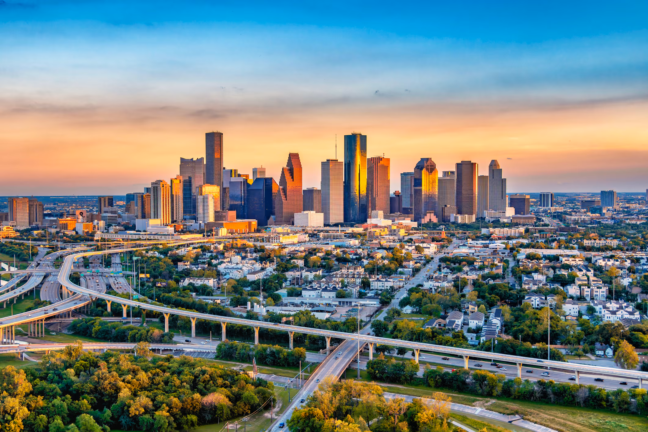 Aerial view of a large city skyline at sunset with tall skyscrapers reflecting warm light, highways with cars, residential areas, and green trees in the foreground.
