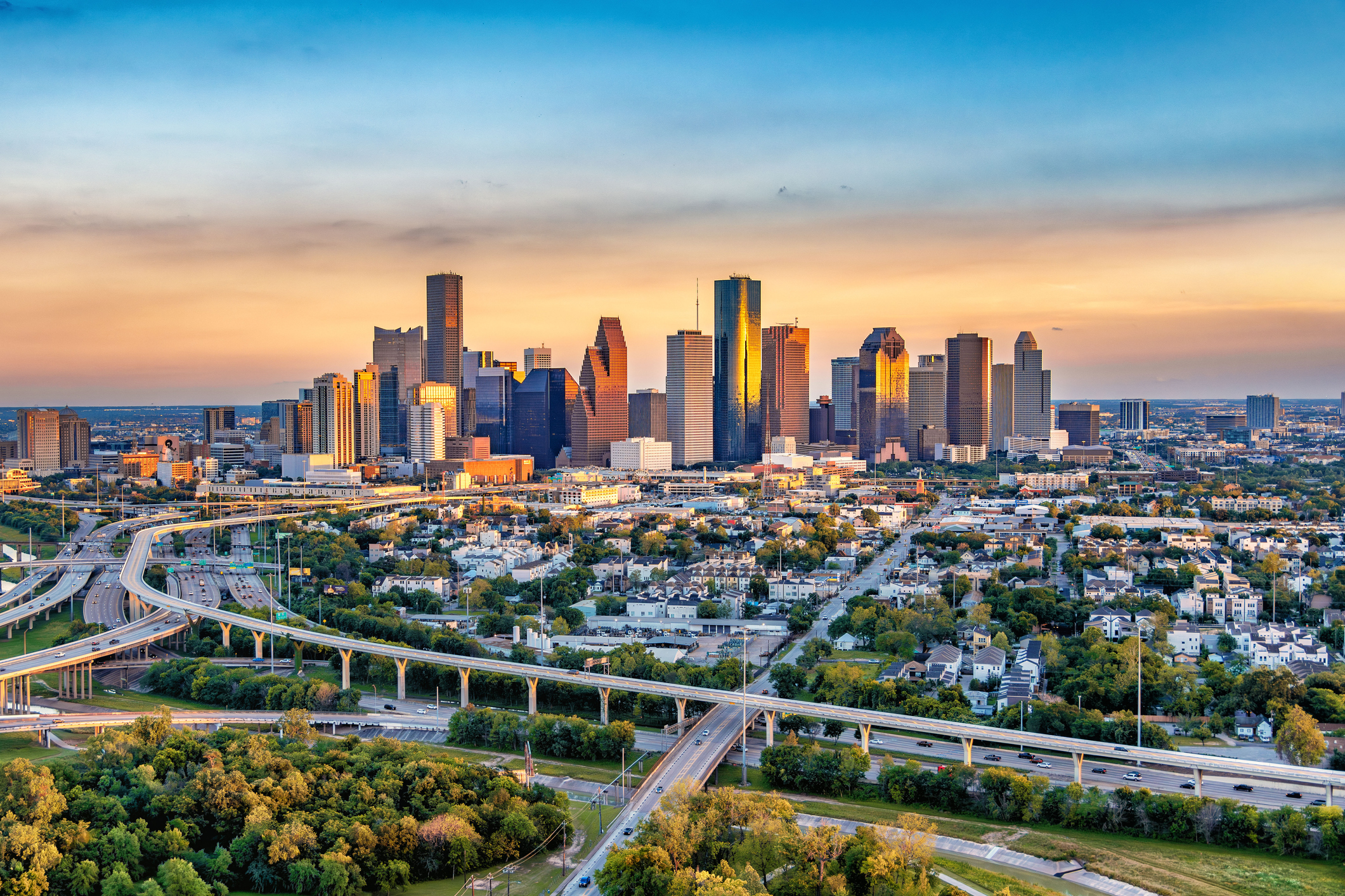 Aerial view of a large city skyline at sunset with tall skyscrapers reflecting warm light, highways with cars, residential areas, and green trees in the foreground.