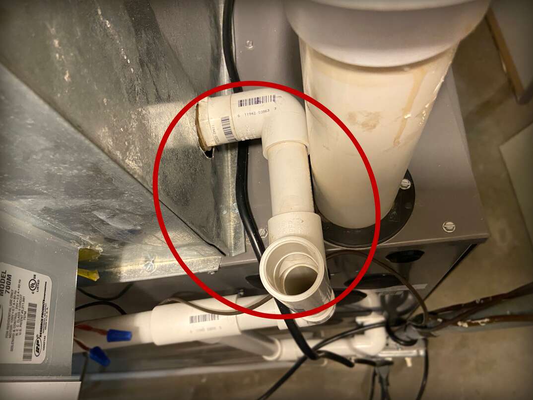 Close-up of white PVC air conditioning drain line pipes under a metal HVAC unit with an open pipe end circled in red.