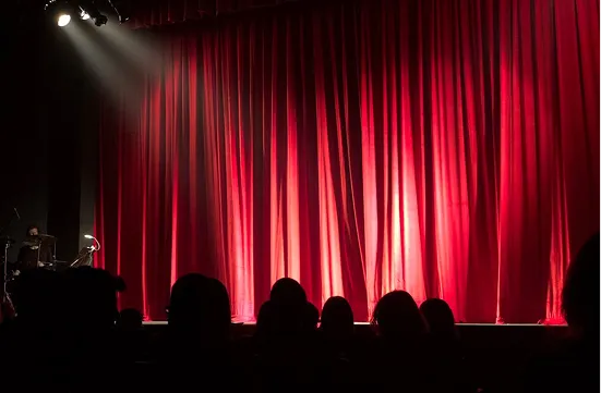 Audience silhouettes facing a brightly lit red theater curtain under stage lights.