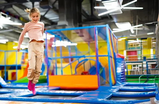 Young girl jumping on an indoor trampoline with colorful play structures in the background.