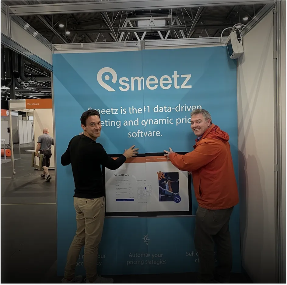 Two men smiling and pointing at a Smeetz booth with marketing and pricing software info.