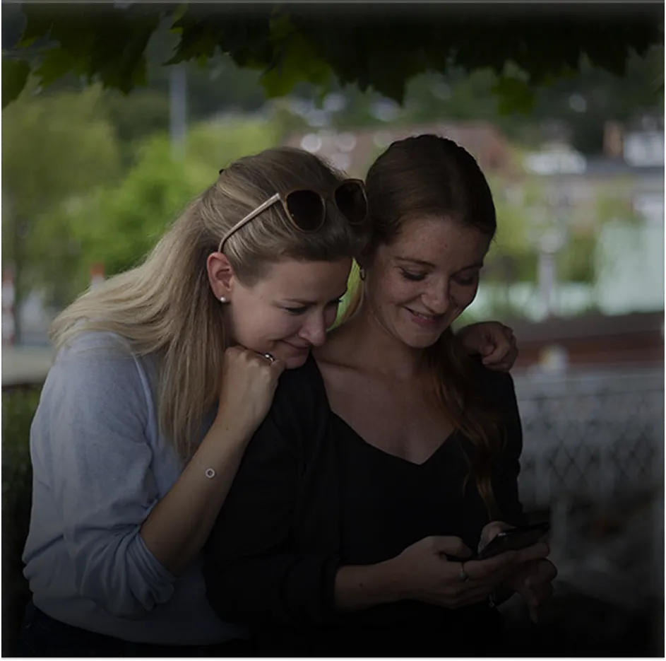 Two women smiling and looking at a phone together outdoors under leafy branches.