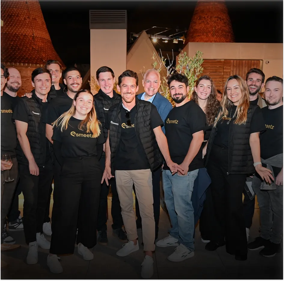 Group of people in black Smeetz shirts smiling and standing together at night outdoors.