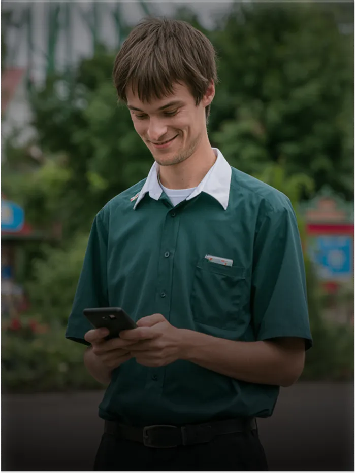 Young man wearing green shirt smiling and looking down at a smartphone in his hands outdoors.