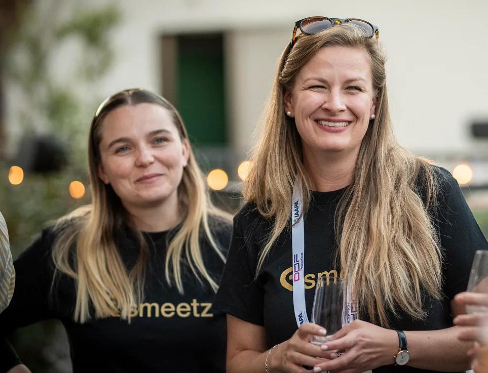 Two women with long blond hair smiling and wearing black shirts at an outdoor event holding glasses.