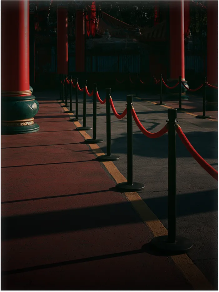 Red velvet ropes on black stanchions forming a queue beside large red pillars in dim light.