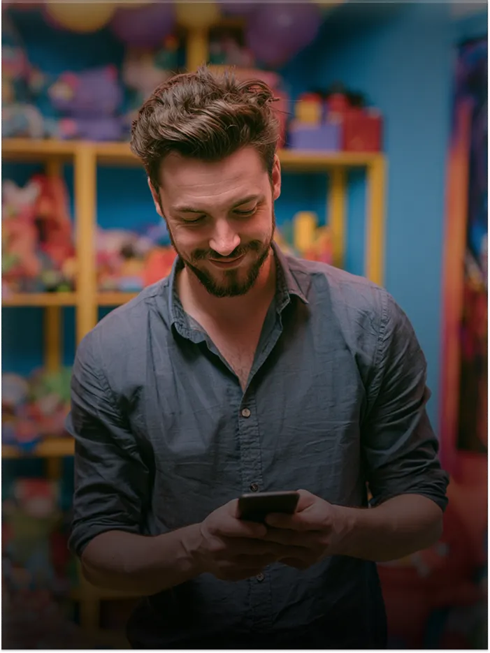 Smiling man looking down at smartphone in a colorful room with shelves of toys.