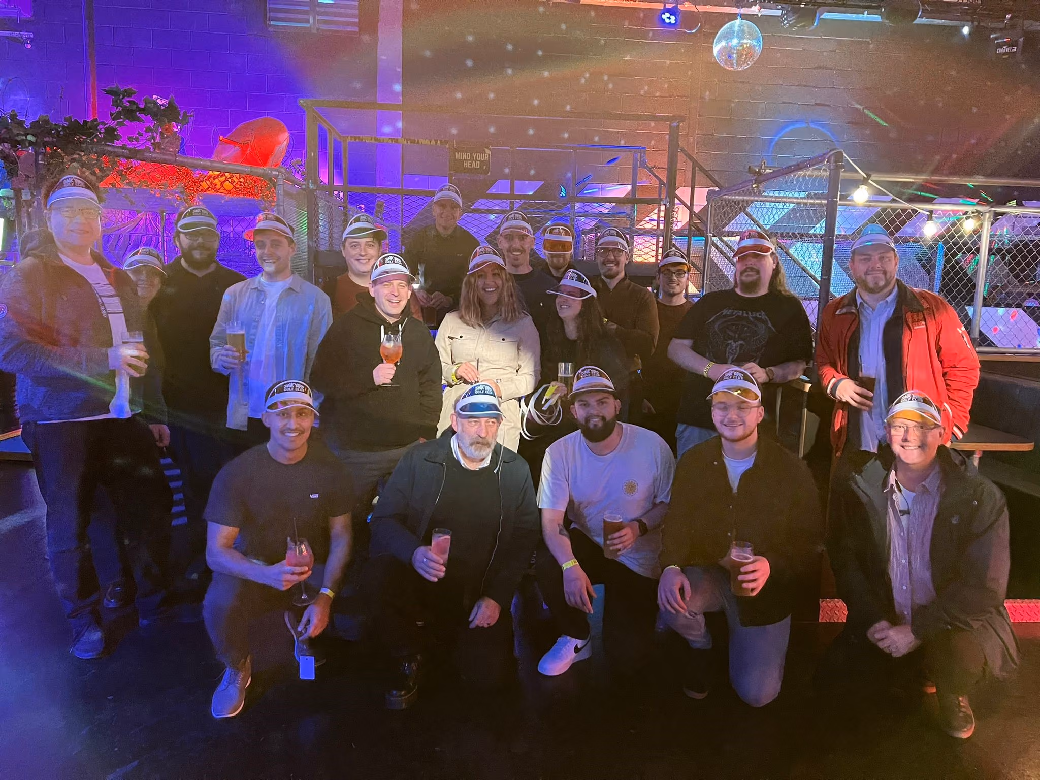 Group of people wearing visors and holding drinks in a dimly lit indoor venue with colorful lights.