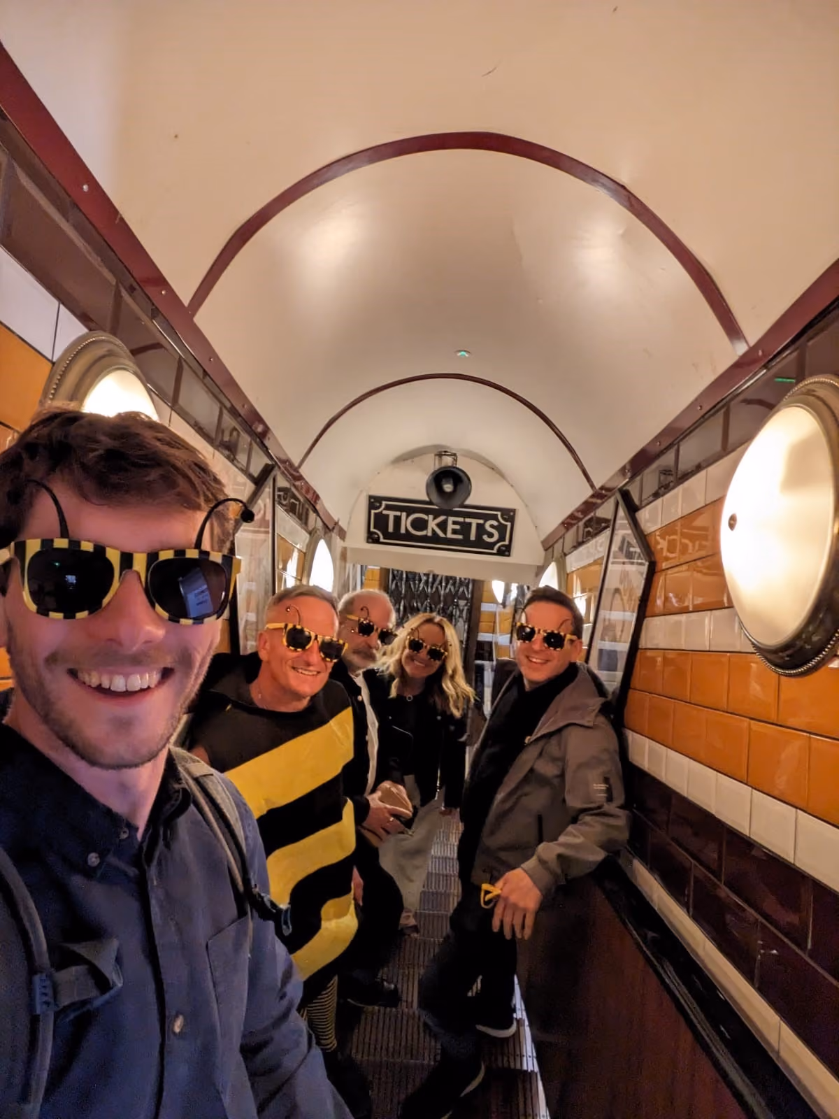Group of five people wearing yellow-black striped sunglasses posing happily in narrow corridor under a tickets sign.