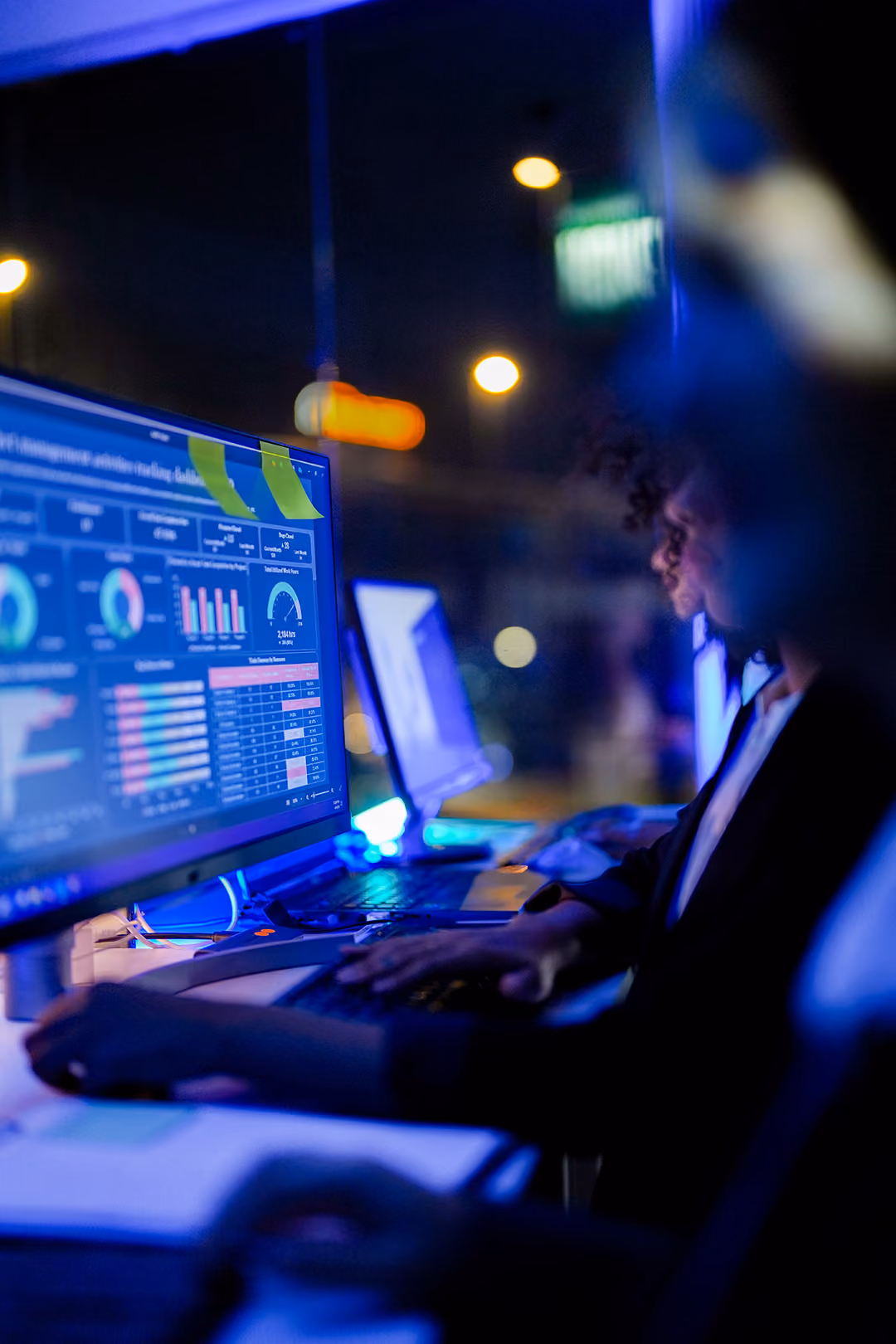 Person using computer at night with data analytics dashboard on screen and blurred city lights outside.