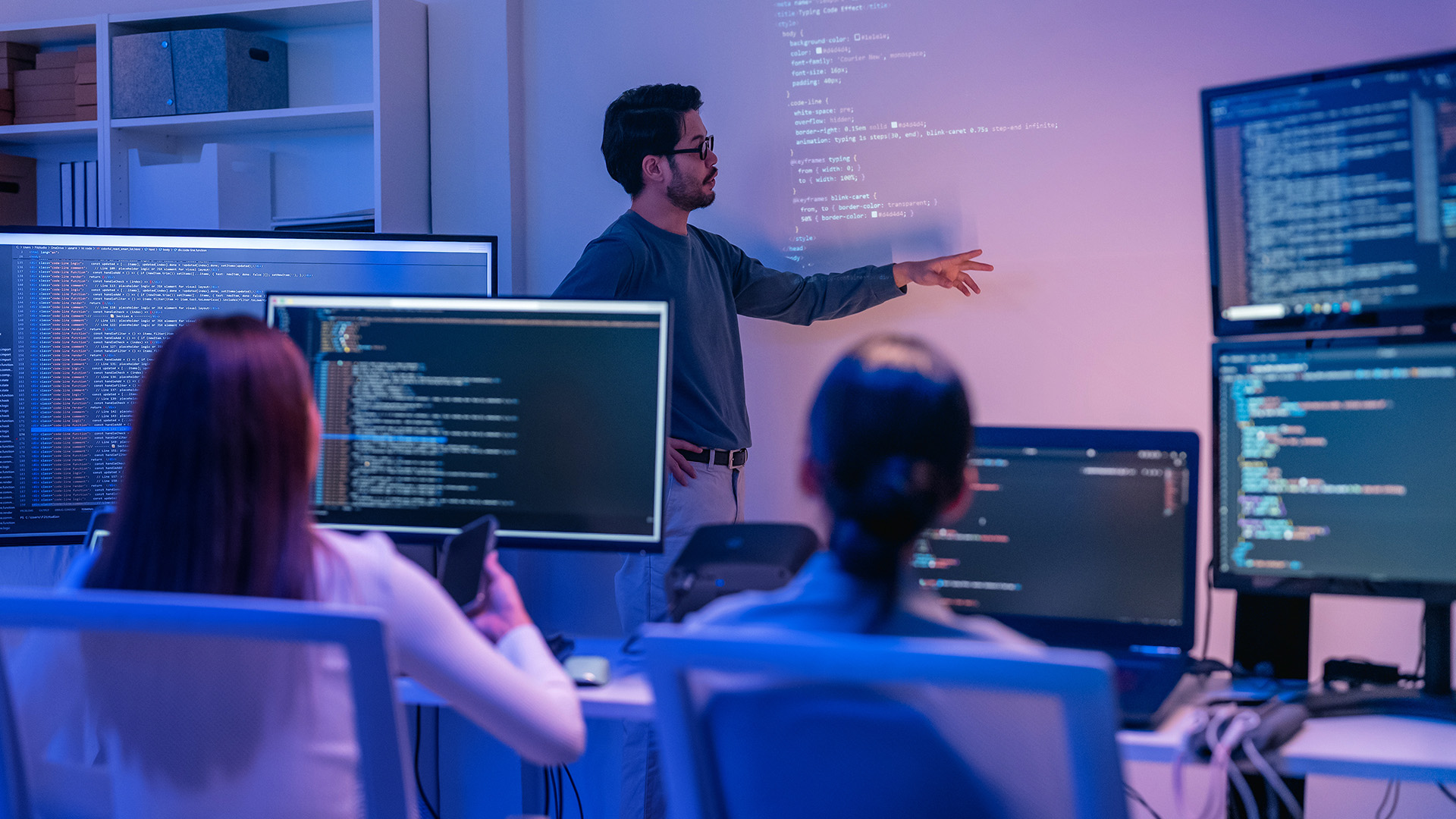 Man presenting software flowchart on screen while three colleagues watch with laptops in a meeting room.