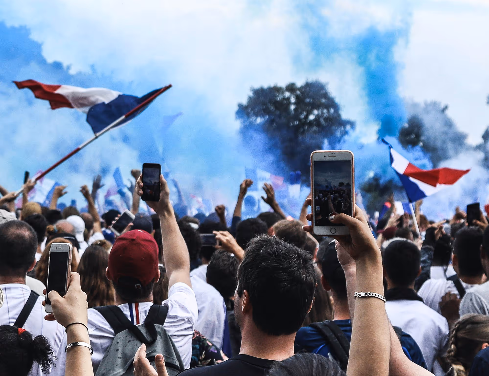 Crowd at an outdoor event holds up smartphones and waves French flags with blue smoke in the background.