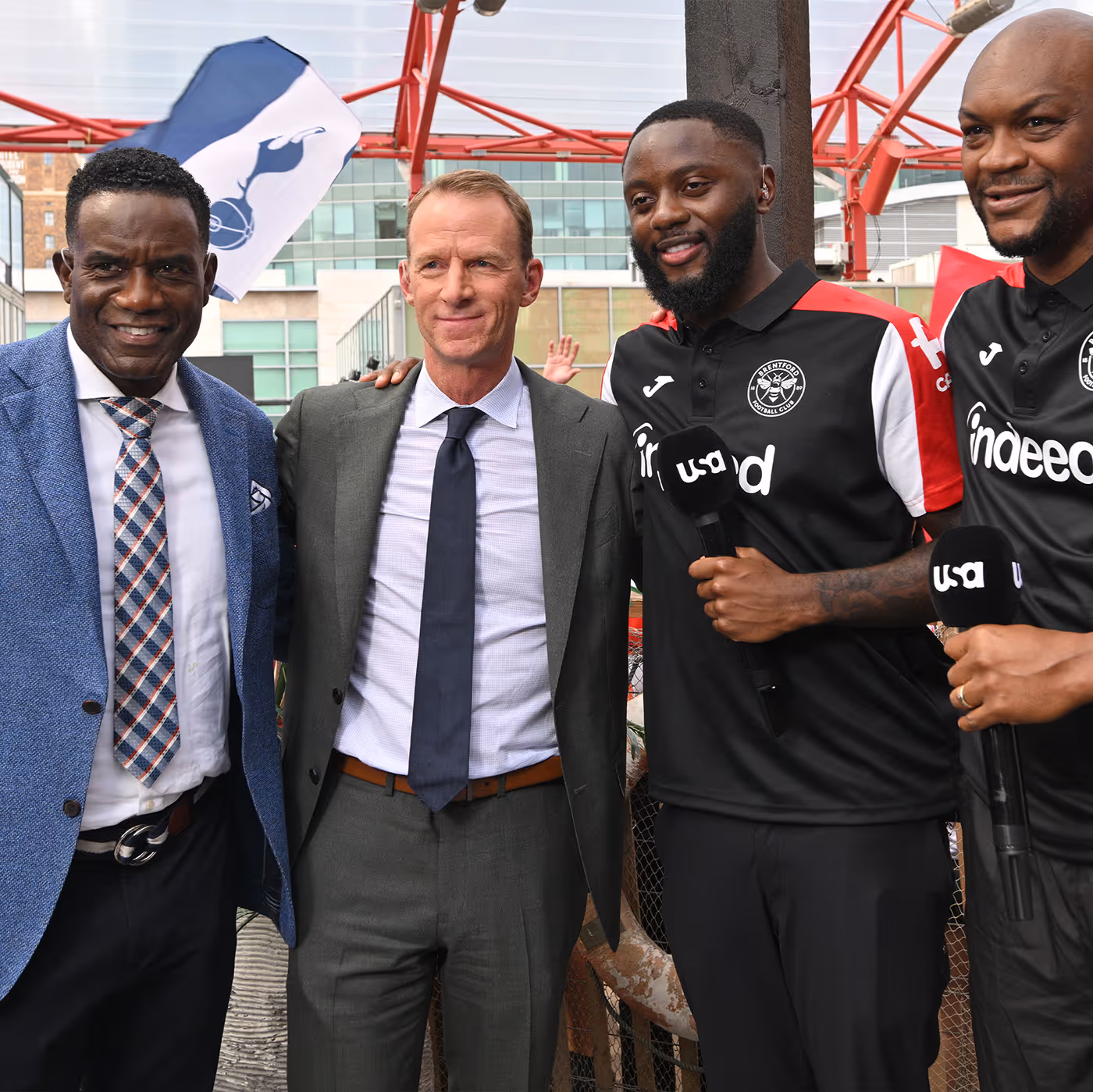 Four men smiling, two in suits and two in Brentford football jerseys holding microphones.