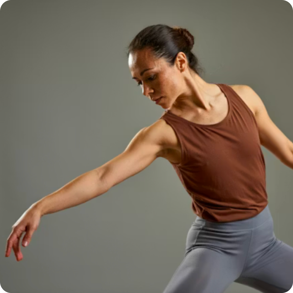 Woman in a brown sleeveless top and gray leggings dancing with extended arms against gray background.