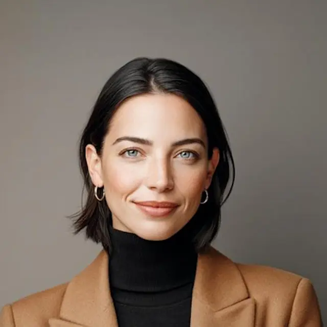Woman with short dark hair and blue eyes wearing hoop earrings, a black turtleneck, and a brown blazer.