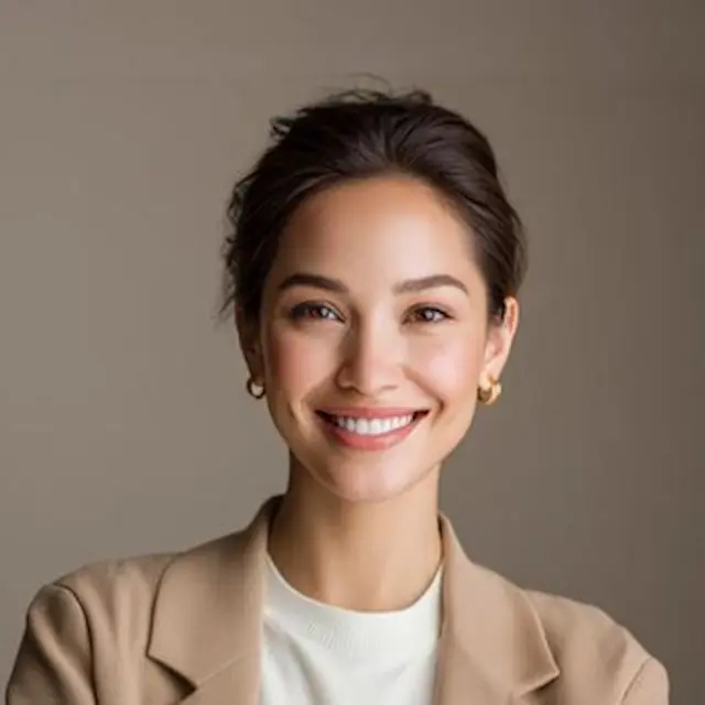 Smiling woman with light makeup wearing gold hoop earrings, beige blazer, and white top on gray background.